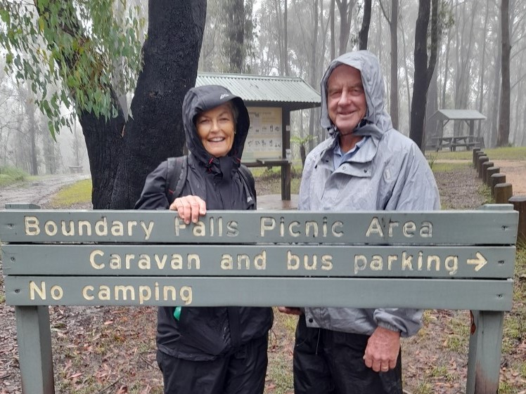Middle aged couple in raincoats outdoors with a sign that says Boundary Falls Picnic Area