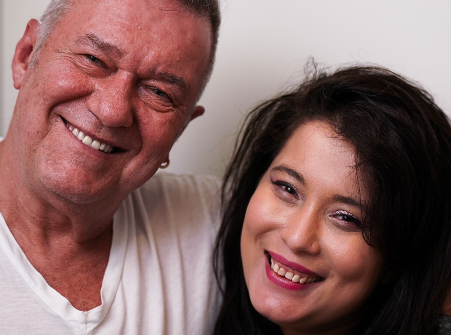 Older man in white T-shirt and woman with black hair and pink lipstick smiling at camera