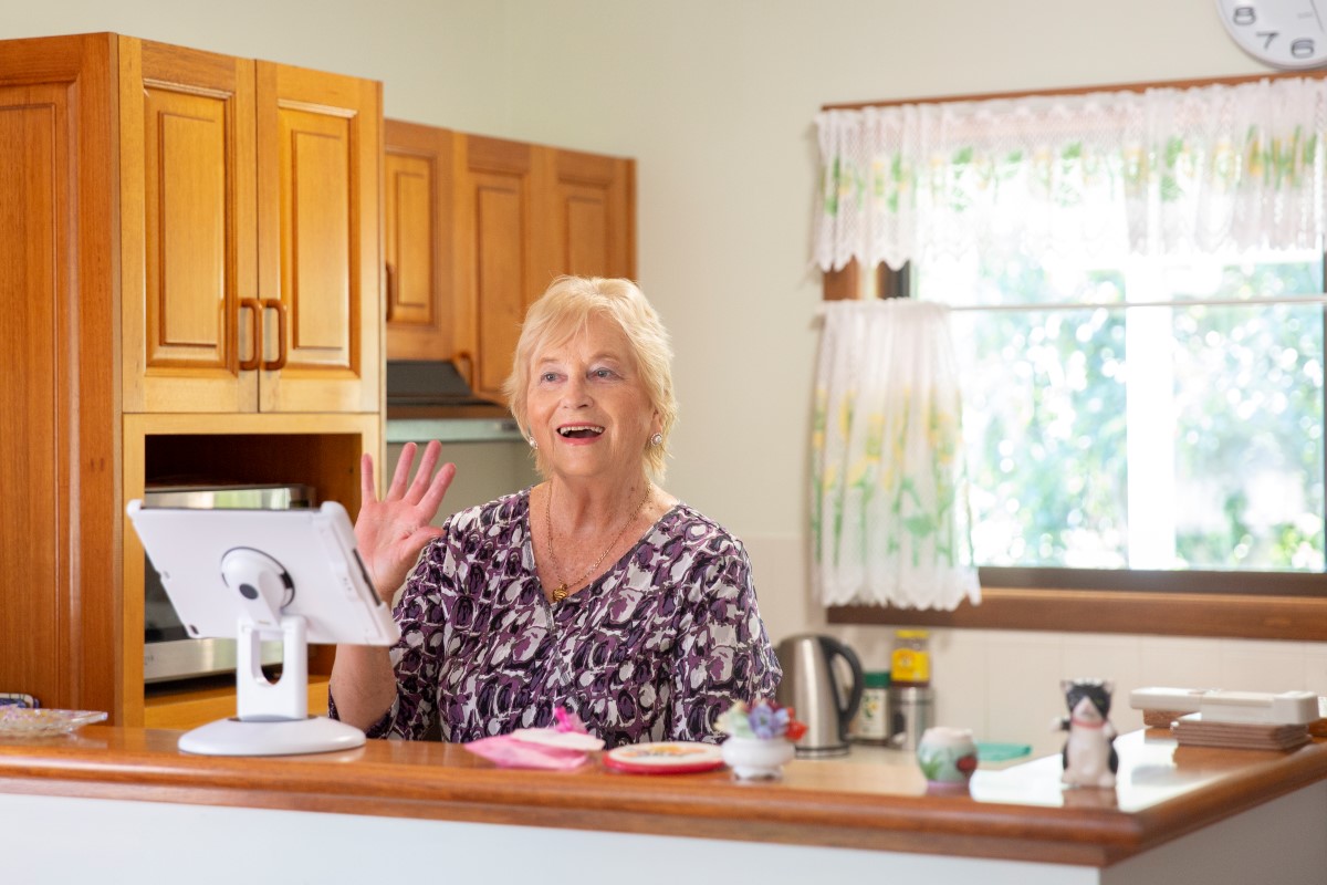 Senior lady in kitchen waving to VSC group on screen 