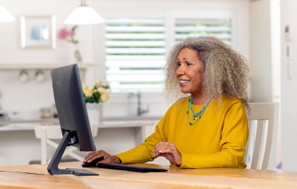 Senior Black lady sitting in front of desktop, wearing bright yellow sweater