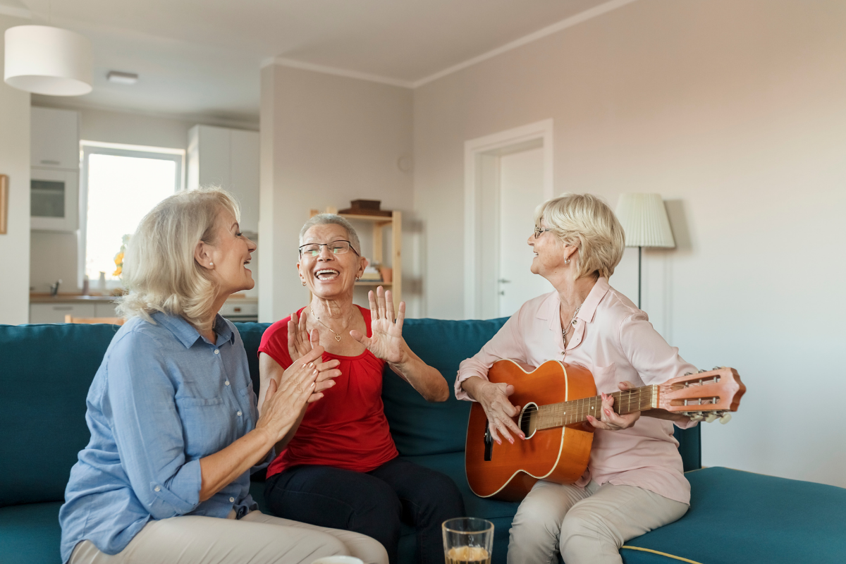 Three senior ladies singing in a living room, one playing the guitar