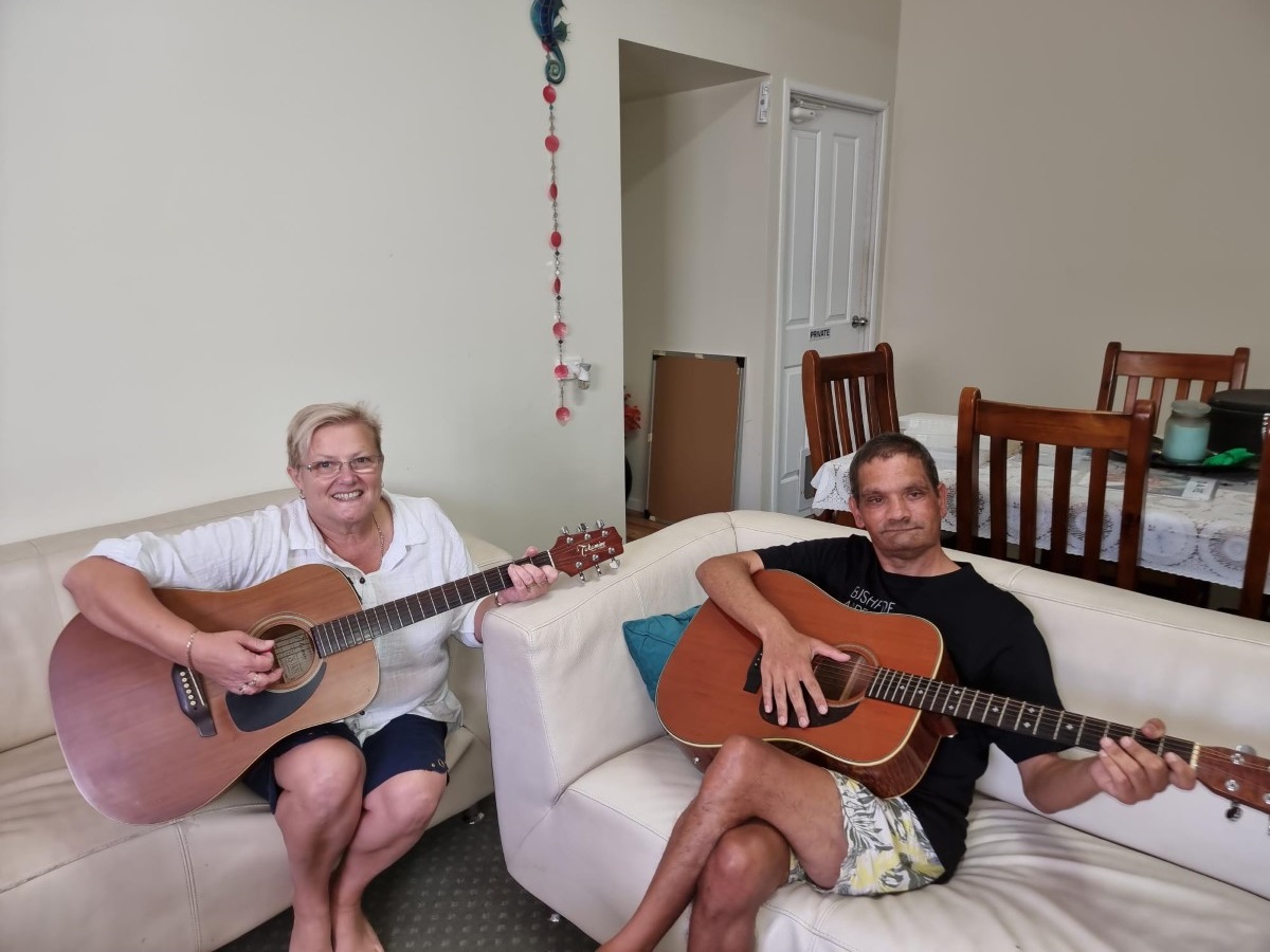 Middle aged blonde woman and man each holding a guitar, sitting on two couches
