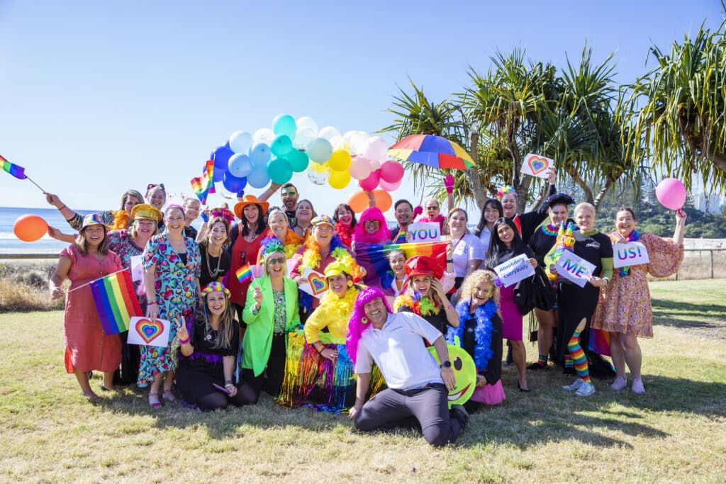 A group of Feros Care employees after the 2022 PRIDE March at Coolangatta Beach. The group is dressed in the various colours of the Pride Flag and are looking at the camera.