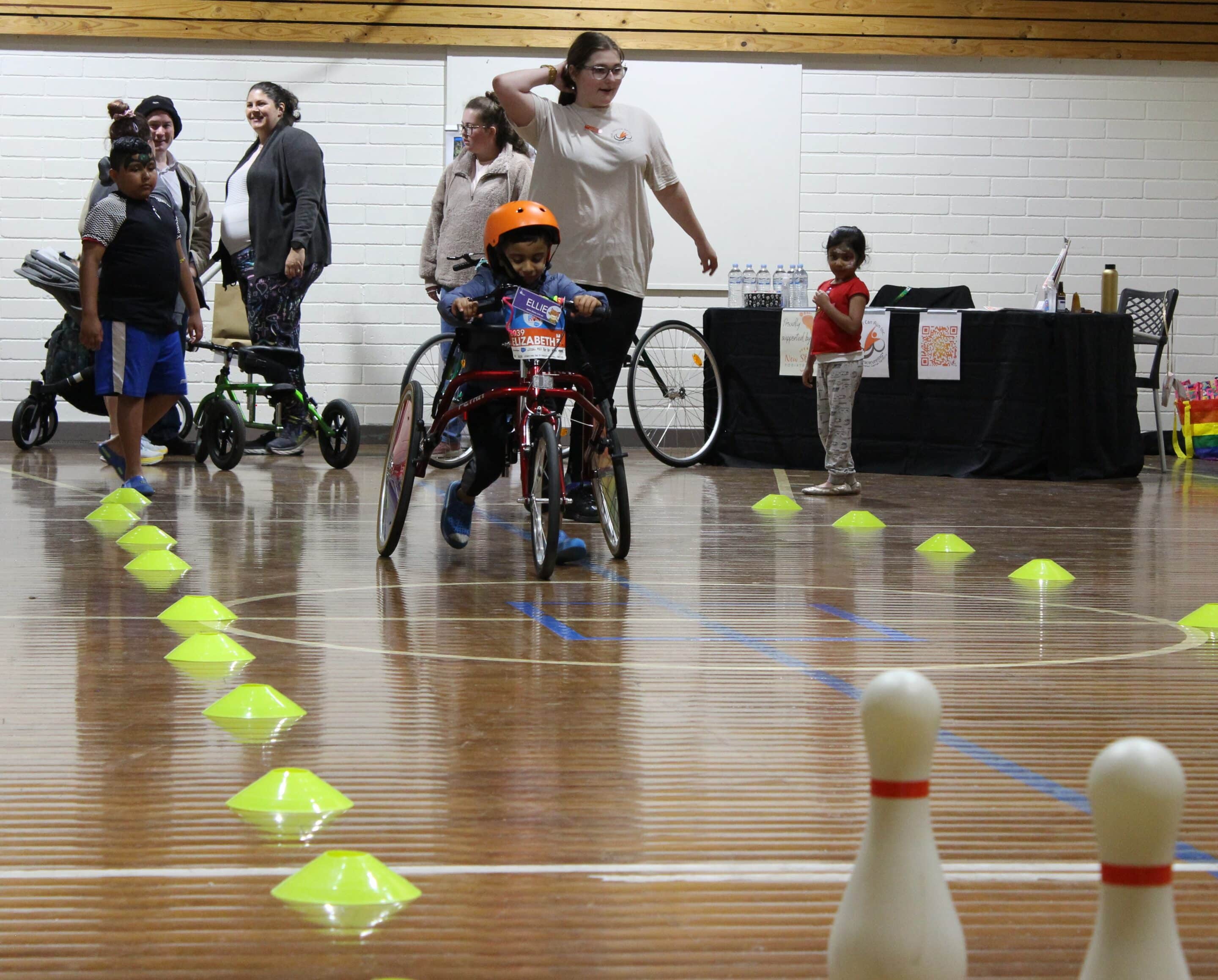 Kids on disability bikes playing ten-pin-bowling indoors at the Accessible Sports and Recreation Expo