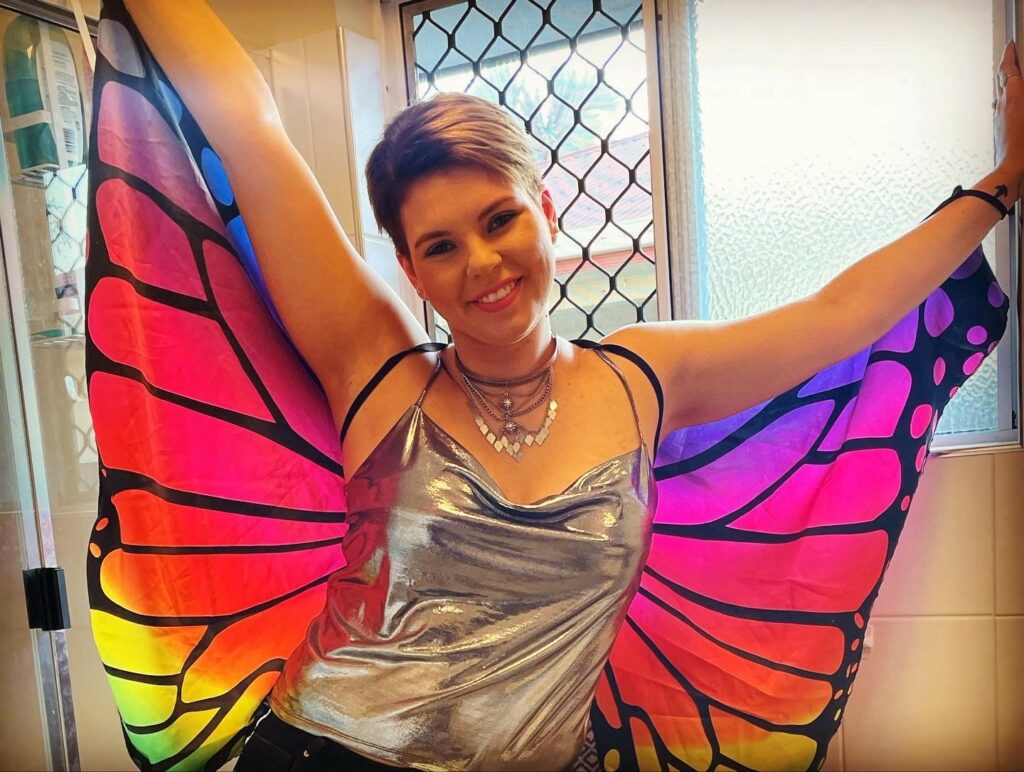 This photo is of Georgia, she is wearing a silver top and bright coloured butterfly wings on her back. Both of her arms are lifted so the large butterfly wings are spread open.
