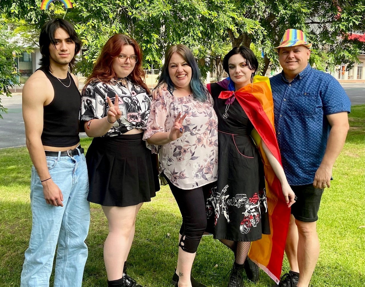 A family of 5 people looking at the camera. One is wearing a Pride flag as a cape, another has a Pride coloured hat on.