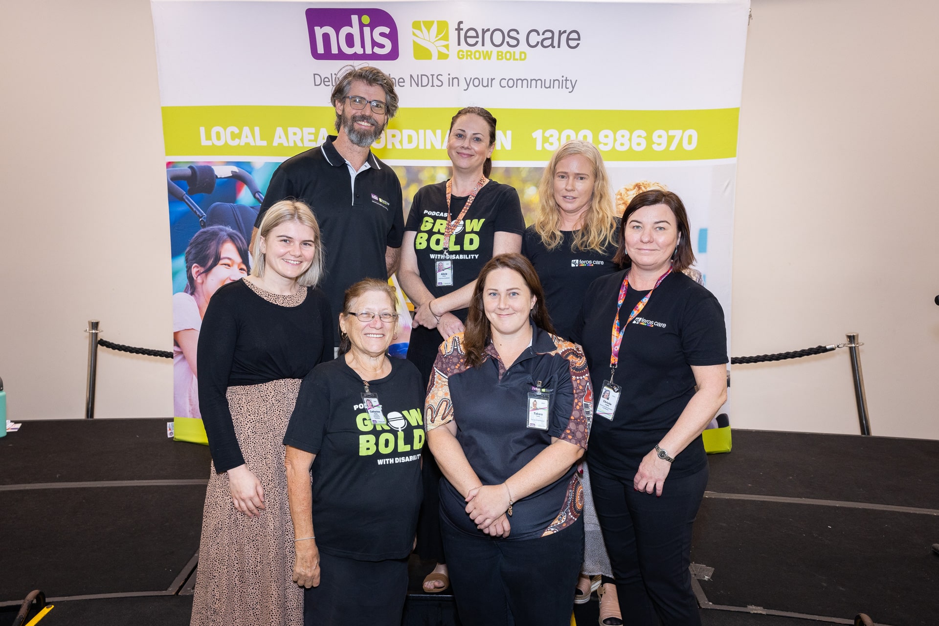 Seven people from Feros Care smiling to camera in front of Feros Care and NDIS banner