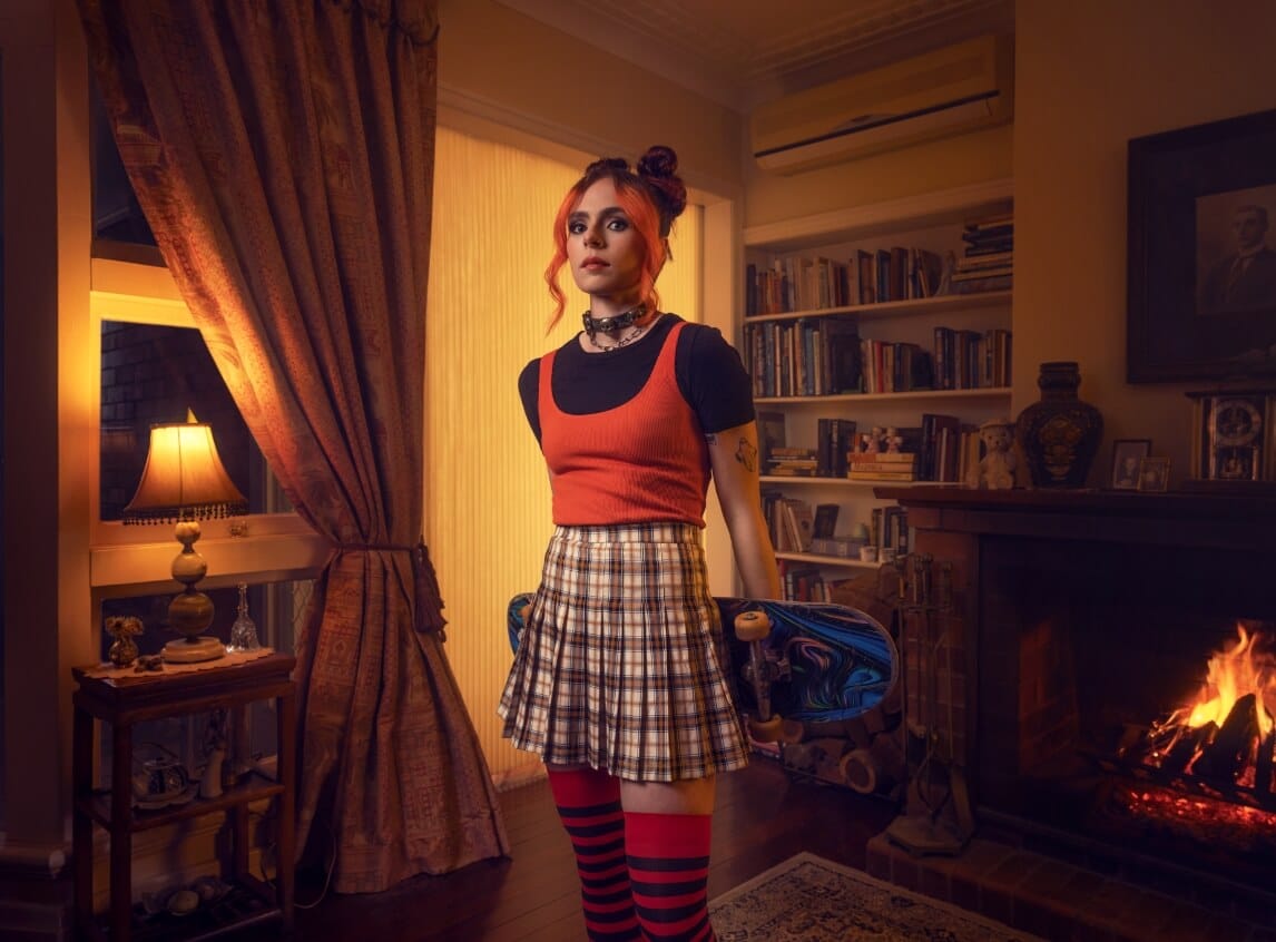 Photo of Zahra, Nursing Assistant at Feros Care, wearing black t-shirt and orange tank top with plaid skirt and thigh length striped socks. Zahra is holding her skateboard and standing in a cozy living room in low light.
