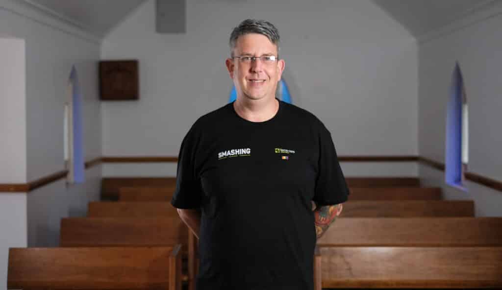 Feros Care Wellbeing Manager Russell Hargreaves standing in church wearing 'SMASHING' documentary series shirt