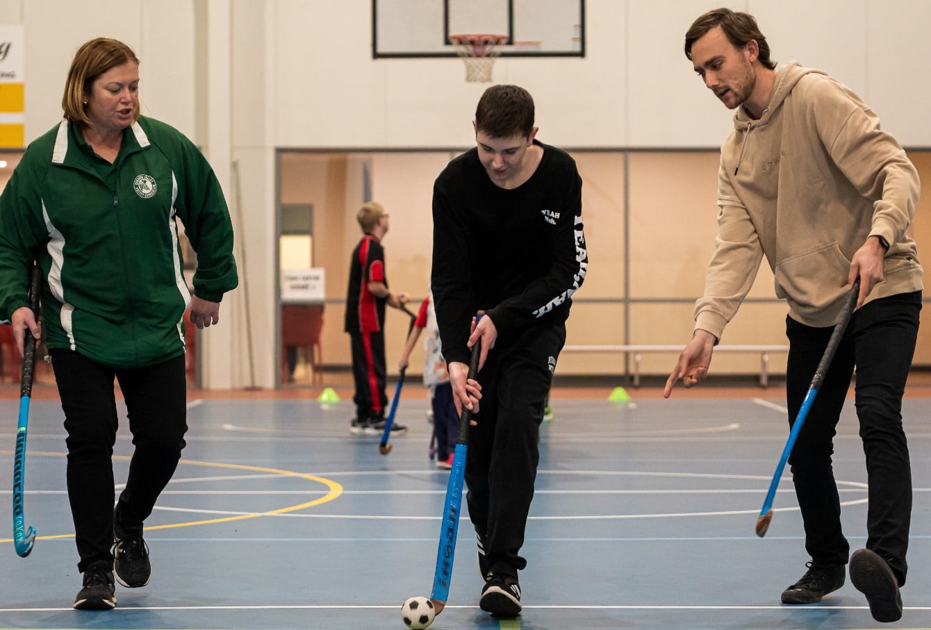 3 people playing a game of accessible hockey. The person in the middle is using his hockey stick to move a bigger than usual hockey ball.