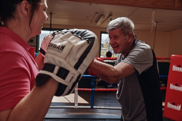Photo of Darryl, Feros Care client, smiling while punching into boxing mitts worn by Kerry, Feros Care physiotherapist