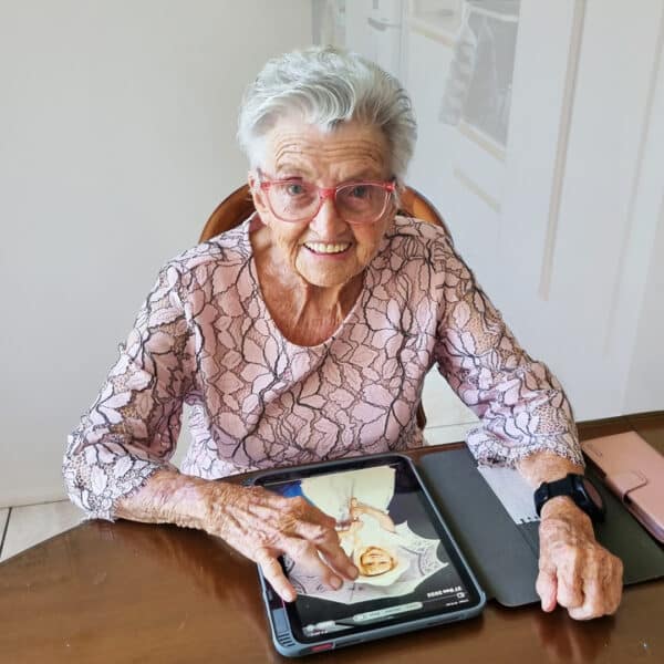 Feros Care client and Let's Get Technical participant Yvonne sitting at her dining room table smiling with her iPad, and wearing her GPS watch.